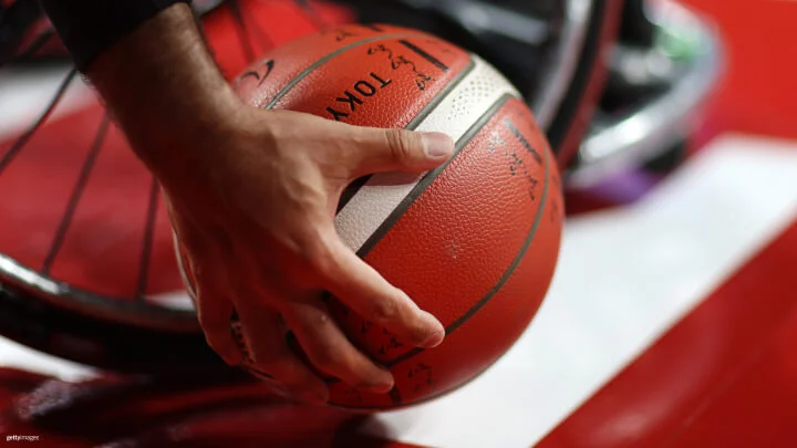 A close-up image shows a hand gripping a basketball. The basketball is orange with black and white lines and has the word "Tokyo" printed on it. The background includes the wheel of a wheelchair, suggesting the setting is related to wheelchair basketball. The focus is on the hand and basketball, with the wheelchair slightly blurred in the background.