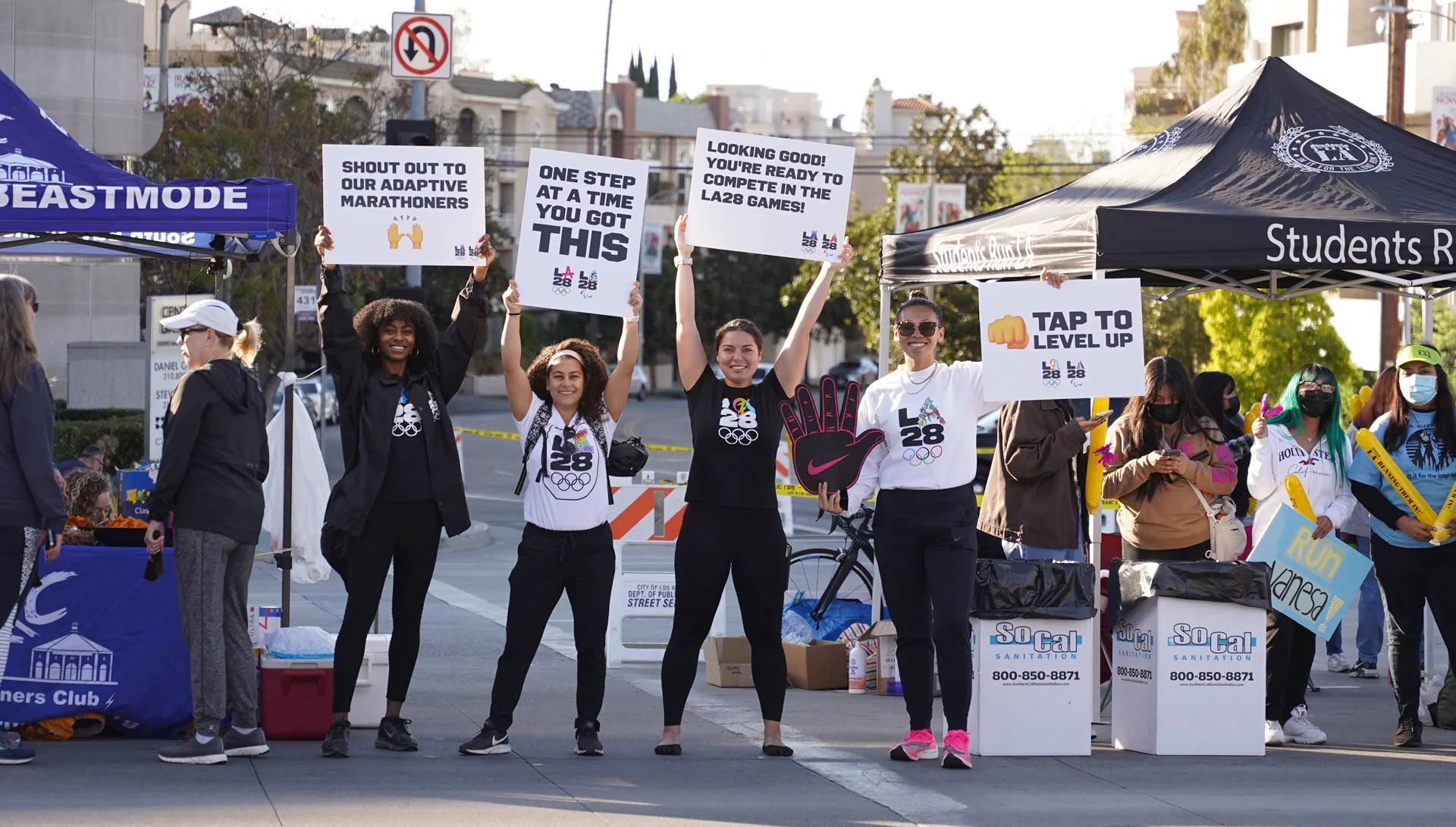 A group photo of four LA28 employees wearing LA28 shirts and holding up signs while standing in a rown among vendor booths with signs that say Shout Out To Our Adaptive Marathoners, One Step At A Time You Got This, Looking Good Youre Ready to Compete in The LA28 Games, Tap To Level Up