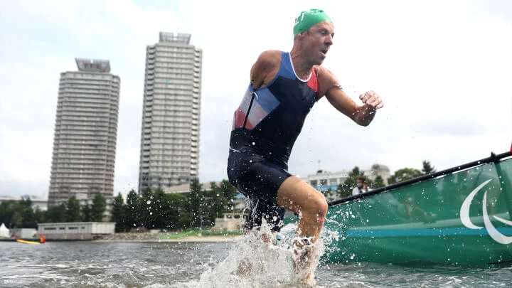 Un atleta Paralímpico, llevando un traje de triatlón y un gorro de natación verde, está emergiendo del agua con gotas salpicando a su alrededor. El atleta tiene amputado el brazo derecho a la altura del hombro. Al fondo, el cielo está nublado y se ven dos edificios altos y algo de vegetación.