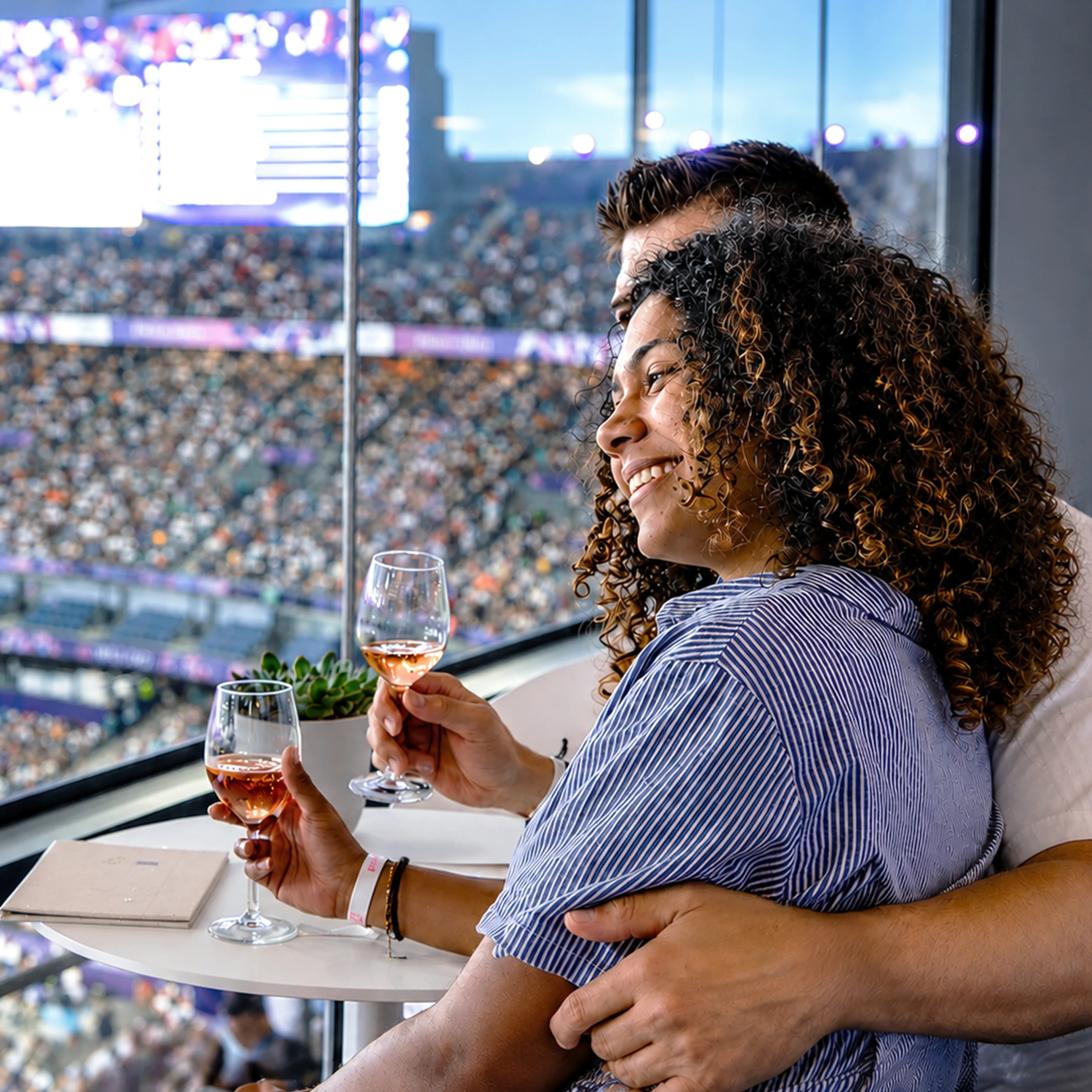 A woman leaning on a man while at the Paris 2024 Games Stade de France Suite while holding glasses of rose wine. 