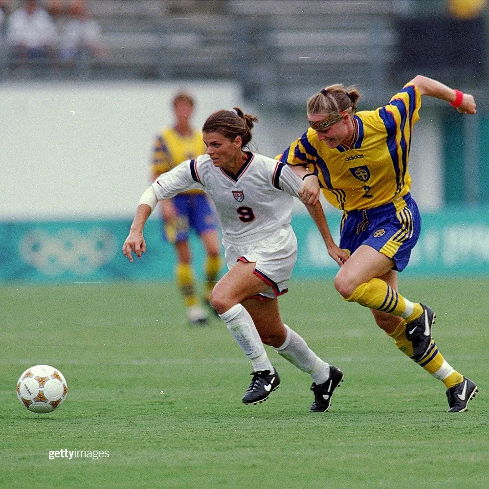 Mia Hamm, integrante de la selección nacional de Fútbol femenino de Estados Unidos, vistiendo medias de Fútbol blancas, tacos Nike, camiseta blanca y pantalones cortos blancos con el número 9, mientras va tras un balón en un campo de césped jugando en los Juegos de Verano de Atlanta 1996 contra Cecilia Sandell de la selección de Suecia, quien viste una bandana estampada, camiseta amarilla y pantalones cortos azules con medias de Fútbol azules y tacos Nike.
