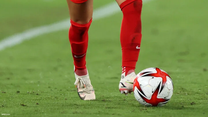 Close-up of a soccer player's legs and feet in motion on a grassy soccer field. The player, wearing red socks and white cleats, dribbles a soccer ball with white, red, and black designs. The grass, slightly worn, indicates recent play.