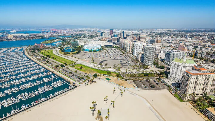 Fotografía aérea del paseo marítimo en Long Beach con un puerto deportivo lleno de botes a la izquierda, una zona de playa arenosa en el centro frontal, un estacionamiento en el centro y numerosos edificios y rascacielos a la derecha, con un cielo azul y vistas de la ciudad en el fondo.