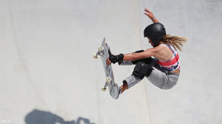 A female skateboarder is captured mid-air performing a trick in a skate park. She is wearing a black helmet, a colorful sleeveless top, grey pants, and protective gear on her wrists and knees. Her long hair is flying back as she holds onto her skateboard, with her knees bent and one hand extended for balance. The background is a smooth, light grey concrete surface.