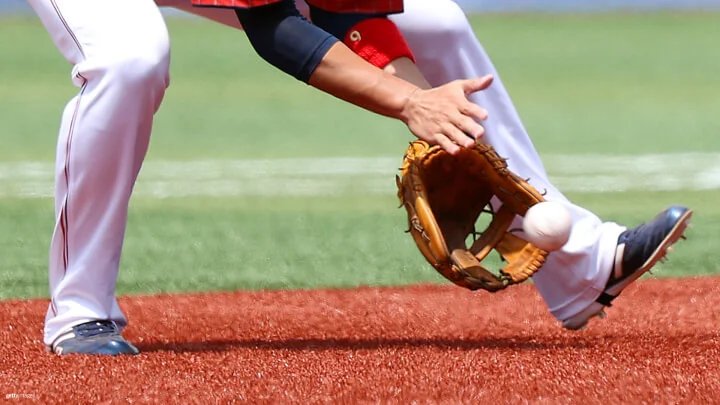 Un jugador de béisbol con pantalones blancos, camiseta roja y tacos negros está en el campo con una pelota rodada. La mano enguantada del jugador está posicionada cerca del suelo a punto de atrapar la pelota mientras que la otra mano del jugador está colocada cerca, lista para asegurar la pelota. El escenario se desarrolla en un campo de béisbol con un césped verde de fondo y un cuadro interior de tono marrón rojizo.