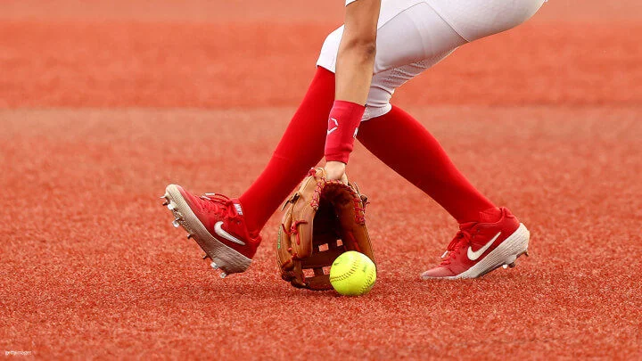 A softball player wearing a dark blue uniform and a white visor, with "USA" written on her jersey, is leaping to catch a yellow ball with her gloved hand. The background shows a blue wall with large white numbers "2020." The player's face is turned upwards, focused on the ball, and her body is stretched out to make the catch.